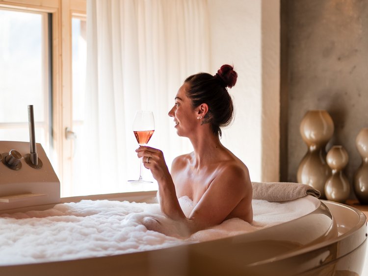 Wie es sich für ein 5-Sterne-Hotel gehört: die Angebote. Frau entspannt im Schaumbad und hält ein Glas Wein in einem gemütlichen Badezimmer