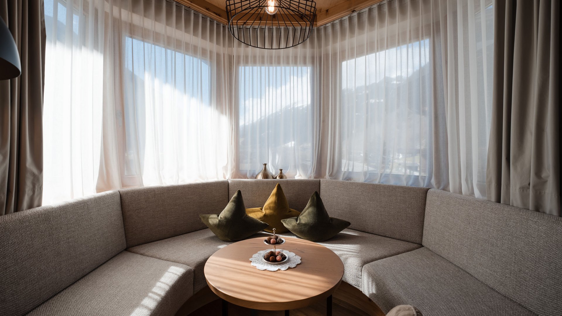 Willkommen im einzigen 5-Sterne-Hotel im Stubaital! Gemütliche Sitzecke mit grauem Sofa und runden Holztisch vor großen Fenstern
