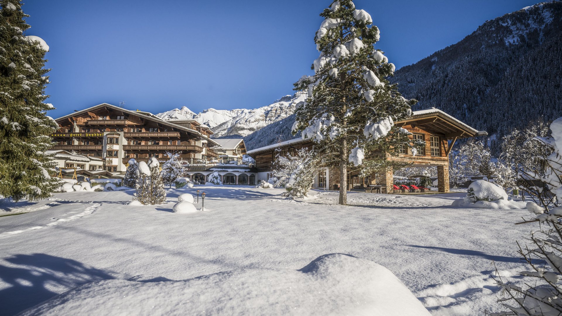 Willkommen im einzigen 5-Sterne-Hotel im Stubaital! Schneebedecktes Chalet und Bäume vor Bergkulisse bei klarem blauem Himmel