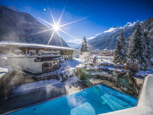 Auf in Ihr De-luxe-Wellnesshotel im Stubaital Winterlandschaft mit beheiztem Außenpool und schneebedeckten Bergen im Sonnenschein
